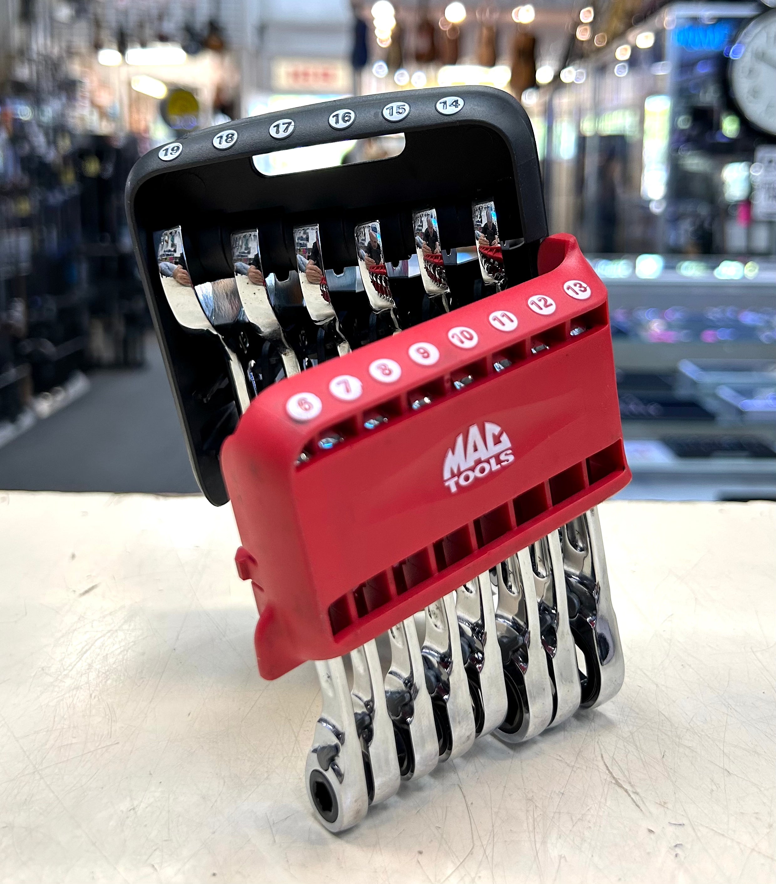 Set of wrenches in a red and black tool holder with 'Mac Tools' branding on a blurred workshop background.